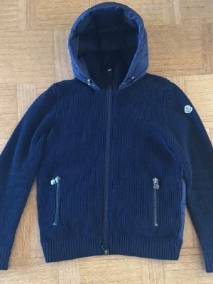 Mörkblå dunjacka från Moncler - Snygg mörkblå dunjacka från Moncler med stickad utsida och quiltad insida. Jackan har huva, två dragkedjeförsedda fickor och Moncler-logga på ärmen. Perfekt för kalla vinterdagar och riktigt skön att bära. Storlek M