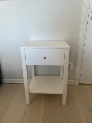 Vit sängbord med låda och hylla - Säljer ett stilrent vitt sängbord i trä med en låda och en öppen hylla undertill. Bordet har raka linjer och en enkel design som passar in i de flesta sovrum. Perfekt för att förvara småsaker och ha böcker eller lampor nära sängen.
