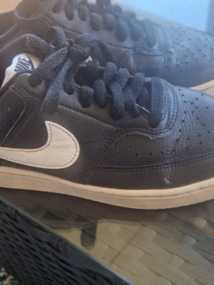 Svarta Nike sneakers med vit sula - Snygga svarta Nike sneakers med vit sula och vit swoosh på sidan. Skorna har klassisk låg profil, svarta skosnören och perforerad tå för extra ventilation. Tillverkade i läder för en stilren look som passar till det mesta.