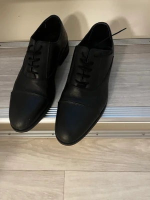 Svarta klassiska loafers i skinn - Stilrena svarta loafers i slätt skinn med rundad tå och diskreta sömmar över tån. Skorna har låg klack och enkel snörning framtill för en tidlös look. Perfekta för dig som gillar minimalistisk och clean stil. Äkta läder på skorna. Pris kan diskuteras😊