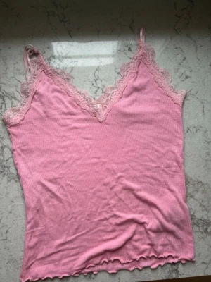 Rosa ribbad linnetopp med spets - Söt rosa linnetopp med tunna axelband och v-ringning. Toppen är ribbad och har fina spetsdetaljer längs urringningen och axelbanden. Perfekt för sommaren och enkel att styla till olika outfits. Storleken är M men funkar för mig som är xs/s!