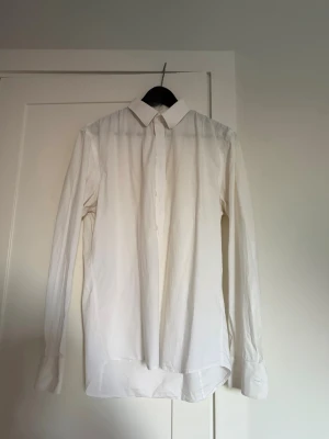 Klassisk vit skjorta med knappar - Säljer en stilren vit skjorta med lång ärm och klassisk krage. Skjortan har knappar framtill och vid ärmsluten, perfekt till en clean och enkel look. Tillverkad i bomull för en skön känsla och bra passform.