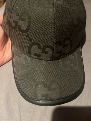 Keps - Snygg svart keps från g.u.cc.i med det klassiska GG-monogrammet i ton-i-ton över hela kepsen. Kepsen har böjd skärm och är tillverkad i ett slitstarkt tygmaterial med detaljer i läder längs skärmens kant. Perfekt accessoar för en trendig streetstyle.