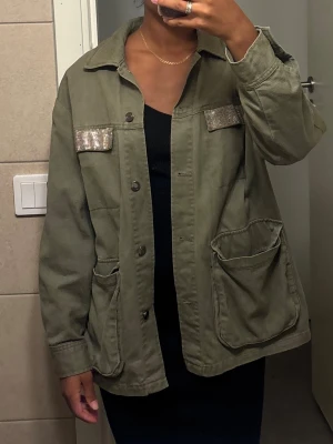 Olivgrön overshirt med paljett-detaljer - Säljer en olivgrön overshirt med stora fickor framtill och snygga paljett-detaljer på bröstet. Jackan har klassisk krage, knäppning framtill och är gjord i ett slitstarkt bomullstyg. Perfekt för dig som gillar en cool och avslappnad stil.