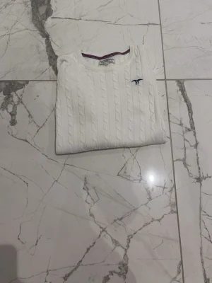Vit kabelstickad tröja Gant - Kabelstickad vit tröja från Gant med rund halsringning och diskret blå logga broderad på bröstet. Tröjan har långa ärmar och ribbade muddar vid ärmslut och nederkant. Klassisk och stilren design som passar till mycket.