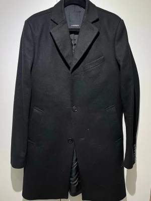 Svart ullkappa från J.Lindeberg - Stilren svart kappa från J.Lindeberg i ullblandning. Klassisk rak modell med två knappar framtill, tre fickor och slät finish. Perfekt för dig som vill ha en tidlös och snygg look till hösten eller vintern.