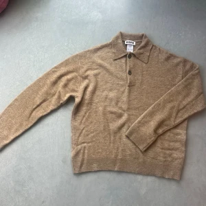 Beige stickad tröja från HOPE - Snygg brun stickad tröja från HOPE med klassisk krage och knäppning framtill. Tröjan har långa ärmar och är tillverkad i mjukt material, perfekt för en stilren och avslappnad look. Tröjan är sliten i designen (se kragen och muddarna). Köpt för någon månad sedan då original priset är 3600kr från HOPE (se bild 4). Aningen croppad och gjord av väldigt fint material. 