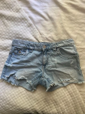 Ljusblå denimshorts - (Storlek 164) Säljer ett par ljusblå denimshorts med råa, fransiga kanter och slitna detaljer. Shortsen har klassisk femficksdesign, bälteshällor och knappgylf. Bakfickorna har snygg broderad detalj. Perfekta för sommardagar!
