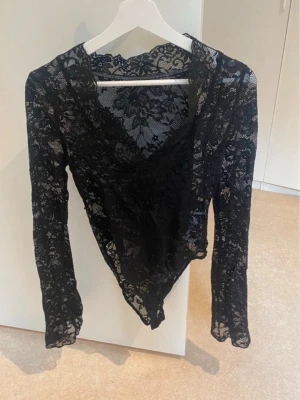 Svart spetsbody med lång ärm - Säljer en svart body i genombruten spets med långa ärmar och djup ringning. Bodyn har ett blommigt spetsmönster och är riktigt snygg att styla till olika looks. Perfekt för dig som gillar trendiga och feminina underkläder. Endast testad