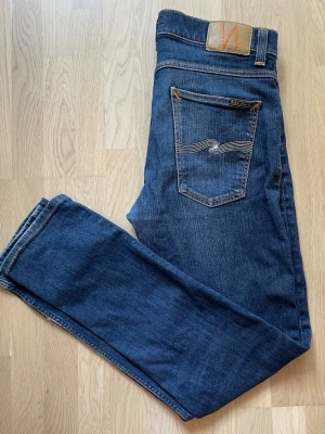 Blå jeans från Nudie Jeans - Säljer ett par klassiska blå jeans från Nudie Jeans med smal passform och snygga kontrastsömmar. Jeansen har fem fickor, läderpatch bak och den ikoniska vågdetaljen på bakfickan. Tillverkade i mjuk denim som sitter skönt hela dagen.