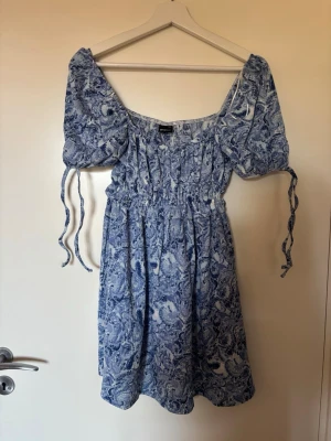 Blåvit kortklänning med puffärm - Söt blåvit kortklänning med puffärmar och knytband. Klänningen har ett blommigt mönster, smock i midjan och en luftig passform. Storleken är XXS men skulle säga den även kan passa XS/S. Helt ny