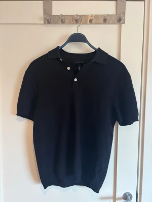 Svart polotopp med kort ärm - Klassisk svart polotopp med korta ärmar och krage. Toppen har knappar framtill och är tillverkad i ett mjukt material som ger en clean och stilren look. Perfekt till jeans eller linnebyxor för en enkel men snygg outfit.