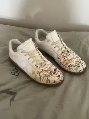 Maison Margiela sneakers med färgstänk - Unika vita sneakers från Maison Margiela med färgstänk i gult, rött, grönt och svart. Skorna har klassisk låg siluett, vita snören och gummisula. Tillverkade i läder med en konstnärlig touch som sticker ut. Bra bra skick 