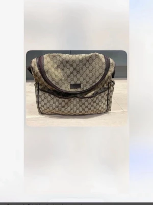 Beige skötväska/axelväska från Gucci - Säljer en beige skötväska/axelväska från Gucci med det klassiska GG-mönstret över hela väskan. Väskan har rundad lockdesign, bruna detaljer och flera fack. Tillverkad i canvas med läderdetaljer. Perfekt för dig som gillar lyxiga accessoarer.