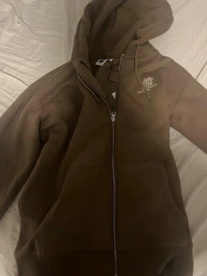 Brun hoodie med dragkedja från Junkyard - Snygg brun hoodie från Junkyard med dragkedja framtill och två stora fickor. Har huva med dragsko och liten vit logga på bröstet '. Perfekt för en chill och avslappnad stil. Ny pris 1000kr