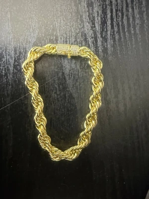 Tjockt guldfärgat rep-armband - Säljer ett chunky armband i guldfärg med tvinnad repdesign. Låset är dekorerat med små gnistrande stenar för extra bling. Perfekt för dig som gillar statement-smycken och vill sticka ut lite extra.