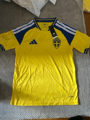 Svensk landslagströja Adidas gul/blå - Gul svensk landslagströja från Adidas med blå detaljer och vita ränder på axlarna. Tröjan har broderat Sverige-emblem på bröstet och Adidas-logga. Tillverkad i lätt och ventilerande material, perfekt för fotboll eller träning.