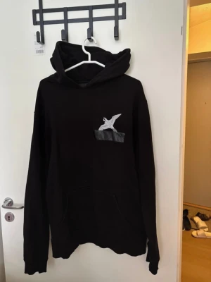 Svart hoodie från Axel Arigato - Snygg svart hoodie från Axel Arigato med huva och en broderad fågel på bröstet ovanför en liten ficka. Hoodien har klassisk passform och är tillverkad i mjukt material som känns skönt mot huden. Perfekt för en avslappnad streetwear-look. Saknar snören. Står ingen storlek på lappen men skulle gissa på att det är en M