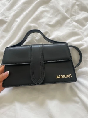 Svart Jacquemus handväska i läder - Snygg svart handväska från Jacquemus i slätt läder med guldfärgad logga på framsidan. Väskan har både topphandtag och avtagbar axelrem, samt en stilren rektangulär form. Perfekt accessoar för att lyfta din outfit.Höjd: 14 cm | Längd: 24 cm | Bredd: 7 cm | aldrig kommit till användning, därmed säljs.