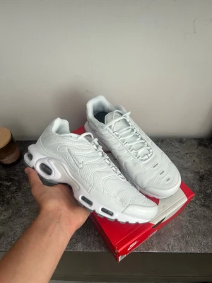 Nike Air Max Plus vita sneakers - Nike Air Max Plus sneakers i helvitt med mesh- och syntetmaterial. Skorna har den ikoniska vågiga designen, synliga Air-enheter i sulan och en liten silverfärgad Swoosh på sidan. Perfekta för dig som gillar stilrena och sportiga sneakers.