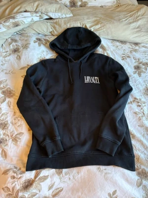 Svart hoodie LOYALTY Unity by Blood - Svart hoodie från LOYALTY med trycket 'LOYALTY UNITY BY BLOOD' på bröstet. Klassisk modell med huva och dragsnören samt en stor magficka. Tillverkad i mjukt material som känns skönt mot huden. Perfekt för en avslappnad och cool stil.