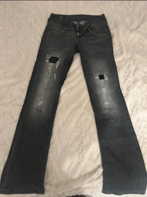 Grå slitna jeans från Lee - Säljer ett par grå jeans från Lee med slitningar och hål på båda benen. Jeansen har klassisk femficksmodell, knappgylf och raka ben. Materialet är denim i bomull och de har en cool, avslappnad vibe.