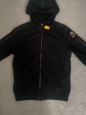 Svart hoodie från Parajumpers - Säljer en svart hoodie från Parajumpers med dragkedja framtill och klassisk huva. På vänster ärm sitter ett stort Parajumpers-märke med texten 'That Others May Live'. Hoodien har ribbade muddar och fickor framtill. Perfekt för en chill och stilren look. Det enda problemet med tröjan är det lilla vid dragkedjan man kan se på bilden annars är allt perfekt och den är 100% äkta.