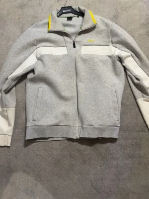 Hugo Boss zip-kofta/tröja strl M (passar även L) - Säljer en snygg Boss zip-kofta i grått med vita detaljer och gul Boss-logga. Mycket fint skick utan hål eller fläckar. Bekväm och stilren modell som passar både till vardags och finare tillfällen.Storlek M (passar även L) ✅ Fint skick ✅ Original Hugo Boss ✅ Dragkedja och fickor