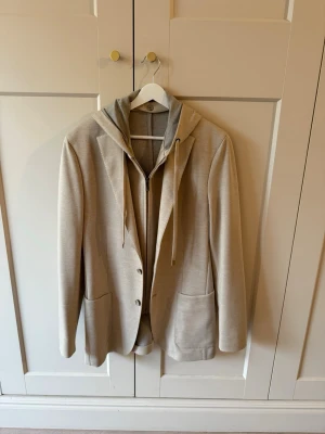 Boggi Madison Sweatshirt Jacket Stretch Cotten blend - Stilren beige kavaj med grå huva och dragkedja, samt snören för en avslappnad streetwear-look. Kavajen har två stora fickor framtill och stängs med knappar. Perfekt för dig som gillar att mixa klassiskt med sportigt. Aldrig använd, extraknappar tillkommer. Nypris 5.690kr