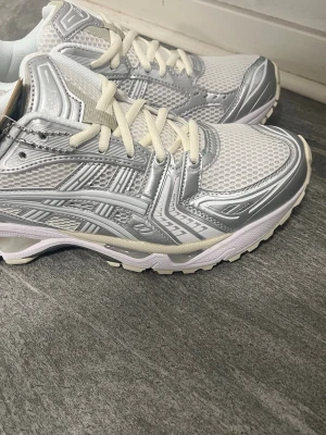 ASICS vita och silver sneakers - Snygga sneakers från ASICS i vitt och silver med mesh-överdel och detaljer i blankt syntetmaterial. Skorna har vita snören och en bekväm, vadderad insida. Perfekta för dig som gillar sportig och modern stil.