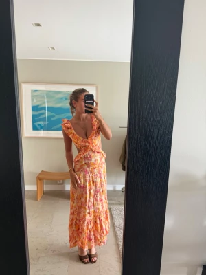 Färgglad långklänning med volang - Säljer en långklänning i härliga orange, rosa och gula toner med volangdetaljer och djup v-ringning. Klänningen har cutouts framtill, öppen rygg och är ärmlös. Perfekt för sommarens alla tillfällen!