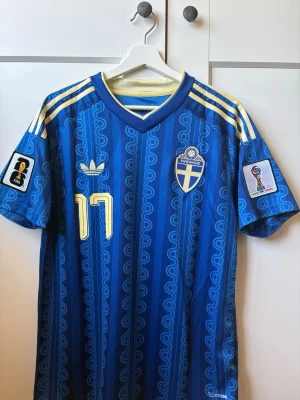 Blå Sverige fotbollströja Adidas - Säljer en blå Sverige fotbollströja från Adidas med gula detaljer och nummer 17. Tröjan har officiella FIFA och VM-märken på ärmarna, samt Gyökeres namn på ryggen. Klassisk v-ringad modell med gula ränder på axlarna och snyggt mönster i tyget. Helt ny, redo för bud!!!