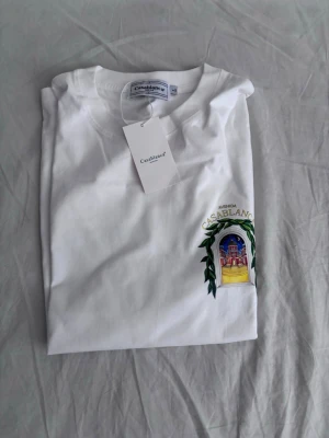  Casablanca t-shirt  - Säljer en vit t-shirt från Casablanca med färgstarkt tryck framtill som föreställer en byggnad och gröna blad. T-shirten har klassisk passform och rund hals. Perfekt för dig som gillar unika prints och stilrena plagg.