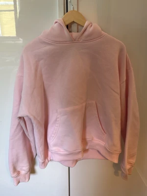 Ljusrosa hoodie med magficka - En oversized ljusrosa hoodie med stor magficka och huva. Hoodien har ribbade muddar vid ärmslut och nederkant. Perfekt för en chill och avslappnad stil. Materialet känns mjukt och skönt mot huden.