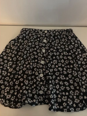 Svart blommig kjol från SHEIN - Supersöt svart kortkjol från SHEIN med vitt blommönster och knappar framtill. Kjolen har resår i midjan för en bekväm passform och är perfekt för dig som gillar en lekfull och trendig stil.