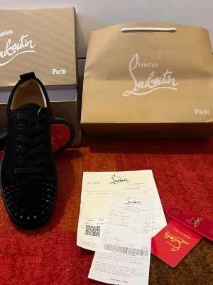 Svarta sneakers från Christian Louboutin - Säljer ett par svarta sneakers från Christian Louboutin med klassisk röd sula och svarta nitar på tån. Skorna är låga, har snörning och är tillverkade i mocka och läder. Snygg och edgy design som sticker ut med exklusiva detaljer.