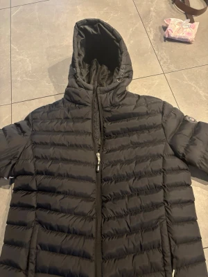 Svart dunjacka från Vinson - Snygg svart dunjacka från Vinson med huva och dragkedja framtill. Jackan har horisontella sömmar och en sportig look, perfekt för kalla vinterdagar. Klassisk design med diskret logga på ärmen och praktiska fickor. Har ett hål under armen