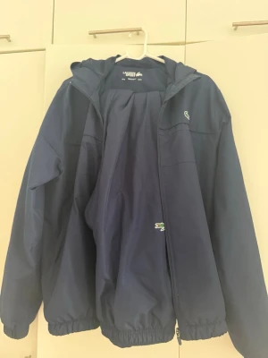 Marinblå träningsdress från Lacoste - Säljer en marinblå träningsdress från Lacoste med jacka och byxor. Jackan har huva, dragkedja och elastiska muddar. Byxorna har resår i midjan och vid benslut. Båda plaggen har den klassiska Lacoste-loggan. Perfekt för sport eller chill.