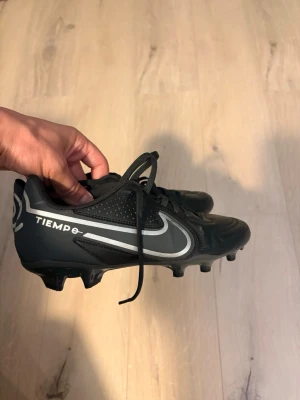Nike Tiempo svart fotbollssko - Säljer ett par helt nya  svarta Nike Tiempo fotbollsskor med vita detaljer och klassisk Swoosh-logga på sidan. Skorna har snörning och dobbar för spel på gräs. Tillverkade i syntetmaterial för bra hållbarhet och passform. Perfekta för dig som vill ha stilrena och funktionella fotbollsskor.