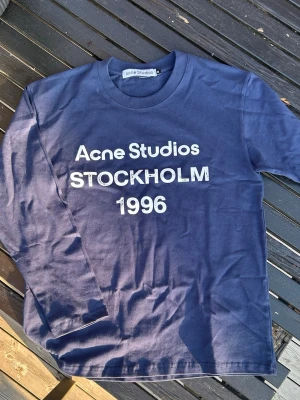 Acne longsleeve blå - Snygg marinblå långärmad t-shirt från Acne Studios med vit text framtill där det står 'Acne Studios STOCKHOLM 1996'. Klassisk rund halsringning och normal passform. Tillverkad i mjuk bomull som känns skön mot huden. Storlek M men passar som en S ish