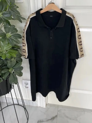 Svart piké med beige loggoband - Svart pikétröja med beige band längs ärmarna, dekorerade med klassiska GG-loggor. Tröjan har krage, knappar framtill och korta ärmar. Perfekt för en trendig och avslappnad stil.