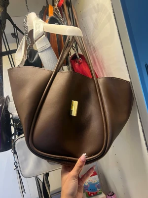 Brun handväska med unik form - Snygg brun handväska i slätt konstläder med unik, vågig form och dubbla handtag. Väskan har ett guldfärgat spänne framtill som detalj. Perfekt för dig som vill sticka ut med din accessoar.