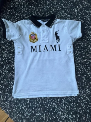 Polo Ralph Lauren - Miami - Vit polotopp från Polo Ralph Lauren med svart krage och ärmslut. Framsidan har broderad logga, emblem och texten MIAMI. Baksidan har en stor svart siffra 5. Klassisk pikélook med knappar vid halsen och detaljer på sidorna.