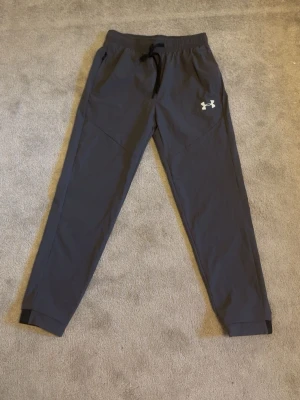 Helt nytt Under Armour Sport joggers, grå, stl M - (Pris kan diskuteras) Snygga mörkgrå mjukisbyxor från Under Armour   med resår och dragsko i midjan. Byxorna har två sidofickor och en vit logga på vänster lår. Tillverkade i ett lätt och mjukt material som ger en avslappnad look.