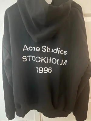 Svart hoodie Acne Studios Stockholm - Svart hoodie från Acne Studios med stor huva och tryck i vitt på ryggen där det står 'Acne Studios STOCKHOLM 1996'. Oversized passform och mjukt material, perfekt för en avslappnad streetwear-look.