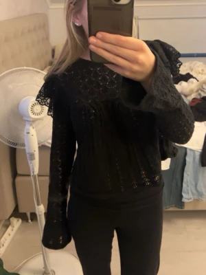 Svart spetsblus med volangdetaljer - Säljer en svart blus i spets med volangdetaljer på axlarna och vida, långa ärmar. 