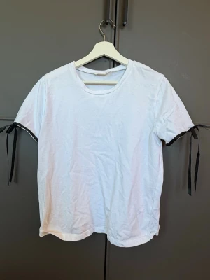 Vit t-shirt med svarta snörningar - Stilren vit t-shirt med korta ärmar och svarta snörningsdetaljer i ärmsluten. T-shirten har en klassisk rund halsringning och är tillverkad i mjukt bomullstyg. Perfekt för en enkel men trendig look.