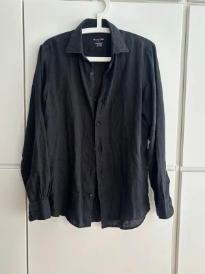 Svart linneskjorta från Massimo Dutti - Stilren svart skjorta från Massimo Dutti i 100% linne. Klassisk krage, knäppning framtill och långa ärmar med manschett. Perfekt för en avslappnad men ändå snygg look. Använd 1 gång.