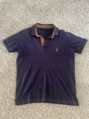 Mörkblå piké från Polo Ralph Lauren - Klassisk mörkblå pikétröja från Polo Ralph Lauren med korta ärmar och kontrasterande brun insida på kragen. Ikoniska loggan broderad på bröstet. Tillverkad i mjuk bomull för en bekväm känsla och snygg passform.
