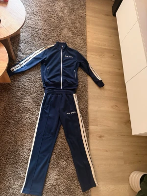 Navy Blå tracksuit från Palm Angels - Palm Angels set i navy blue, storlek M. Mycket fint skick, 9/10. Använt sparsamt och väl omhändertaget utan större anmärkningar. Ett stilrent och eftertraktat streetwear-set med hög komfort och premiumkänsla. Perfekt för både vardag och en mer uppklädd streetwear-look. Säljes då det inte längre används.